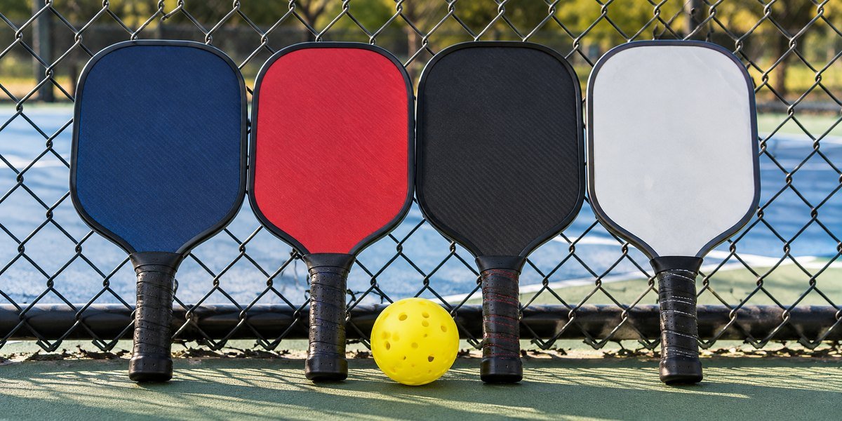 Navy, red, black and white pickleball paddles leaning on a fence with a ball